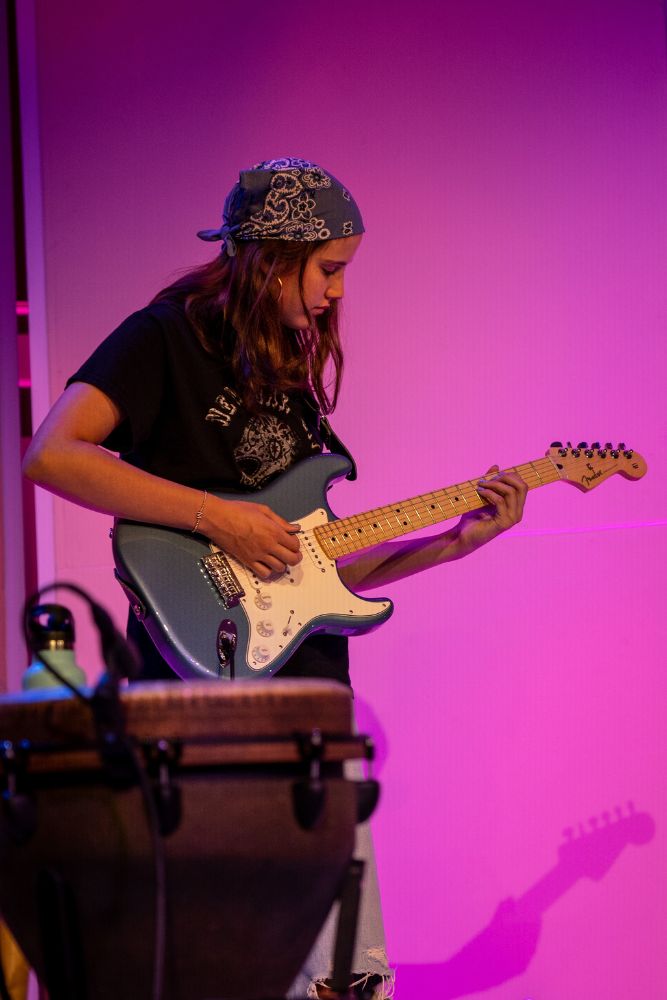 A girl playing guitar on a stage with a florescent background.
